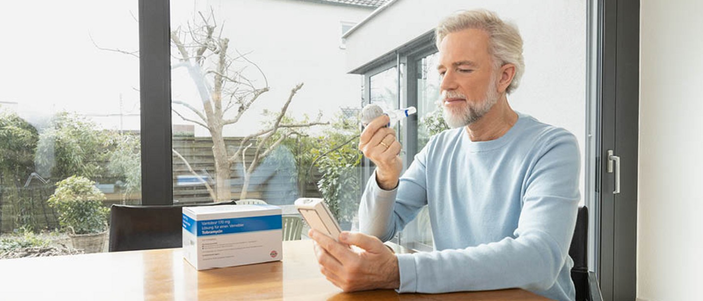 Inhalation for Cystic Fibrosis Woman sitting at a table using a nebuliser connected to an inhalation device while looking at a laptop, as a man stands nearby.