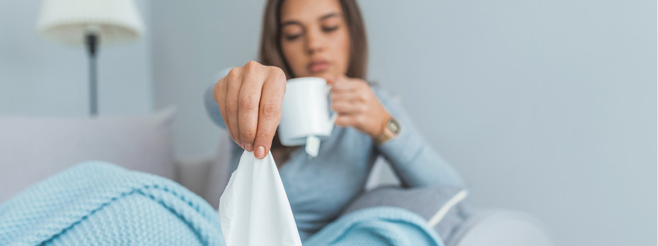Acute or chronic sinusitis? Woman sitting in bed under a blue blanket, holding a tissue and a white mug, appearing unwell or sick.