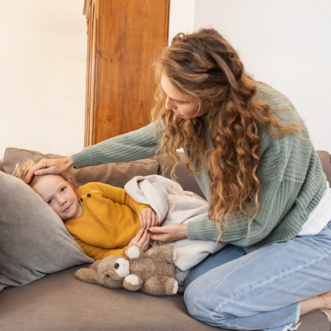 Your child has asthma: What can you do if they catch a cold? Paediatrician Emmanuel Nellen shares his advice Mädchen liegt auf der Couch, Mutter sitzt daneben und hält die Hand auf die Stirn der Tochter