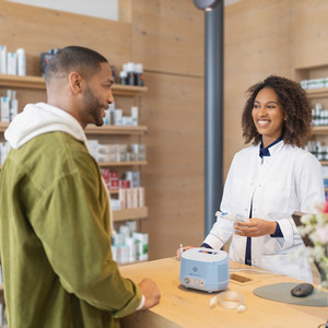 Junger Mann steht vor Tresen in der Apotheke, junge Apothekerin erklärt im das PARI COMPACT2 Inhalationsgerät