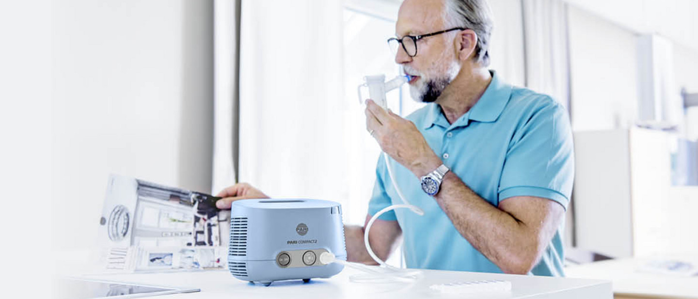 Inhalation for cough and bronchitis Man in a light blue shirt using a nebuliser connected to an inhalation device on a white counter while holding a magazine.
