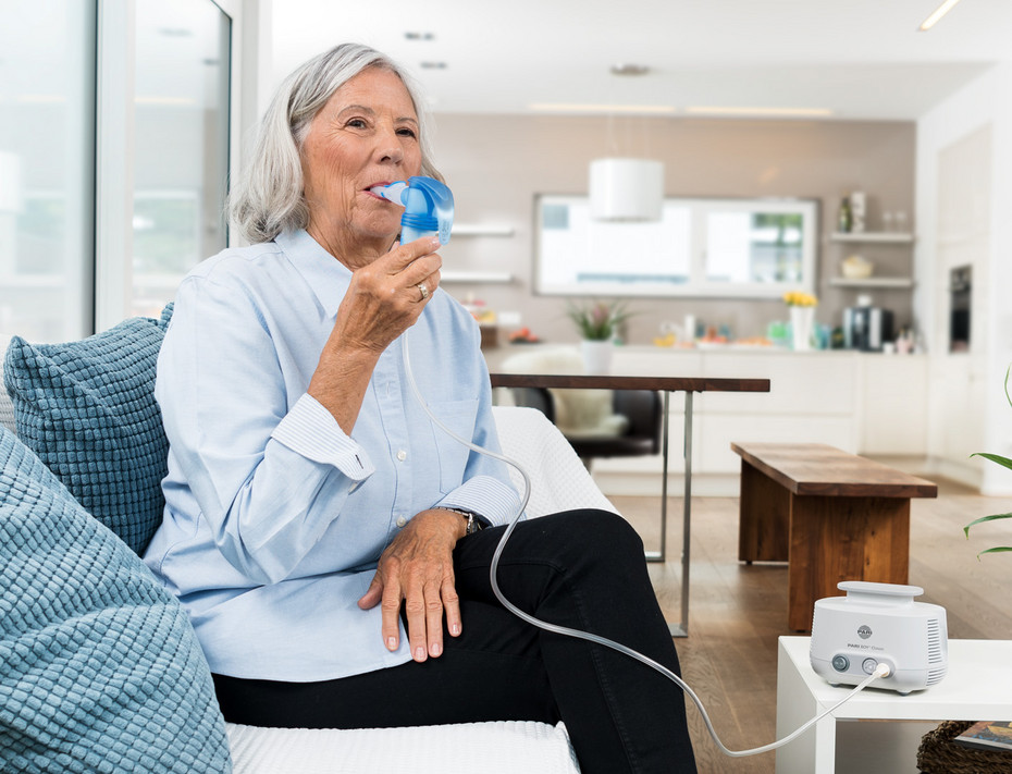 An elderly woman sits relaxed on a sofa and uses a PARI LC SPRINT nebulizer An elderly woman sits relaxed on a sofa in a living room. She uses a PARI LC SPRINT nebulizer connected via a tube to the PARI COMPACT2 compressor on the table.