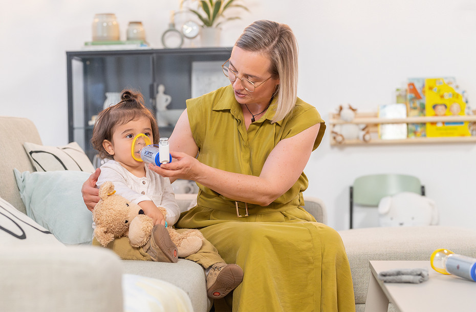Correct use of asthma spray A mother is holding an asthma inhaler with a VORTEX inhalation aid and a yellow baby mask over her toddler’s nose and mouth on the sofa.