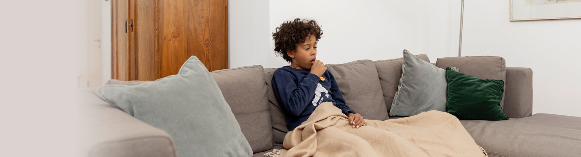 A sick boy sits at home on the sofa under a blanket and has to cough