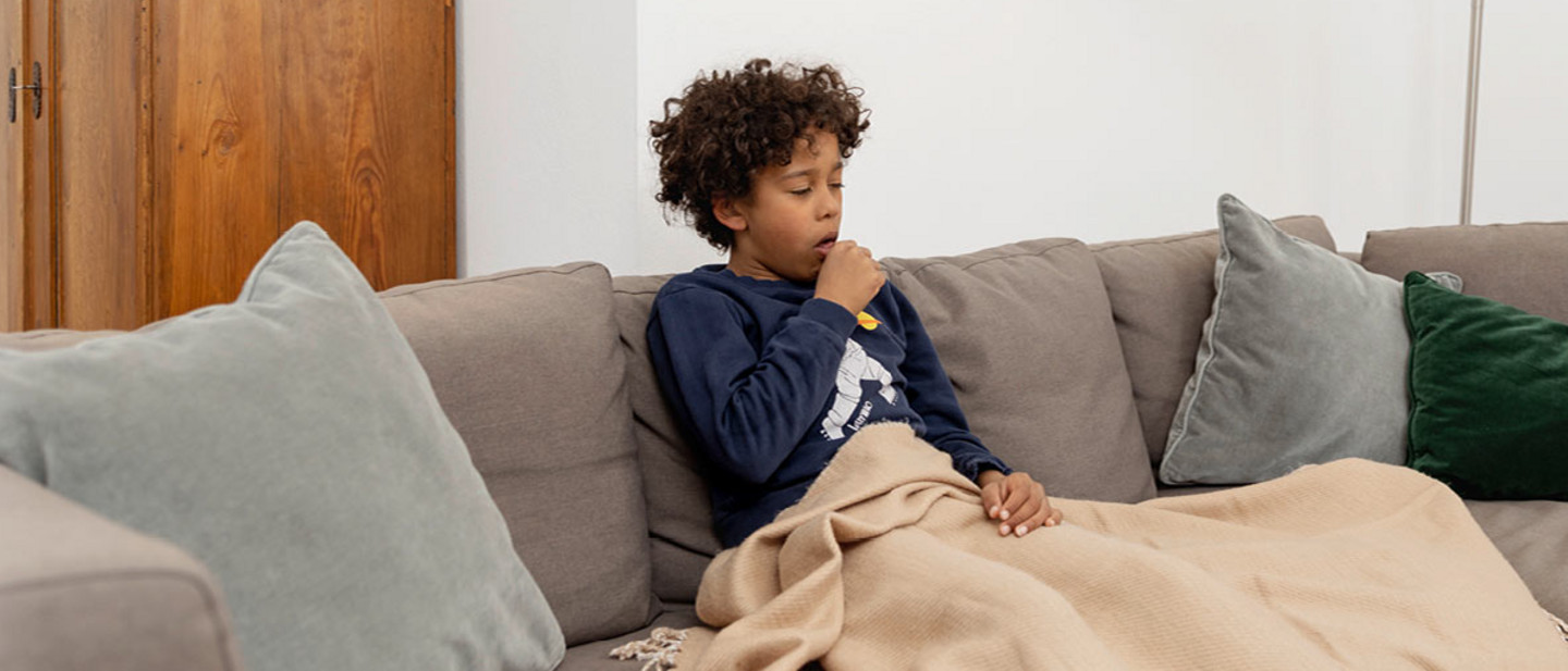 Treatment of RSV Boy sitting on a sofa with a blanket over the legs, raising a hand to the mouth in a coughing gesture.