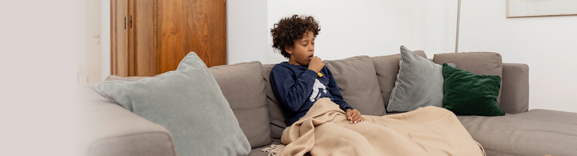 A sick boy sits at home on the sofa under a blanket and has to cough