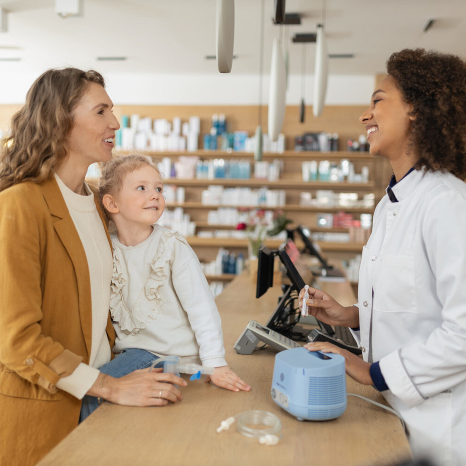 Husten mit Aufwurf: Feuchtinhalation mit einem Vernebler wie dem PARI COMPACT2 Junge Mutter steht vor Apothekerin an der Theke, Kind sitzt auf der Theke, daneben steht ein PARI COMPACT2 Inhalationsgerät