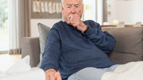 A cough with sputum – what causes the annoying mucus and what to do about it Älterer Mann sitzt hustend auf Sofa, eine Hand als Faust vor dem Mund, die andere Hand greift nach einem Taschentuch
