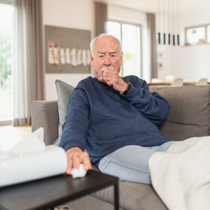 Elderly man sitting on sofa coughing, one hand as a fist in front of his mouth, the other hand reaching for a handkerchief