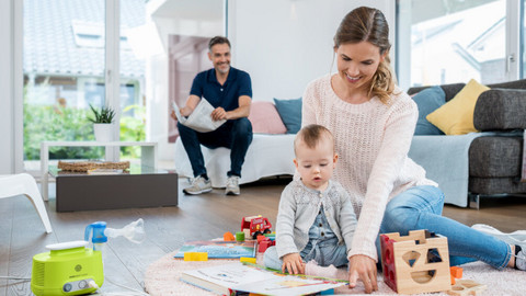 Hypertonic saline solution is suitable for children and infants Mutter sitzt mit Kleinkind auf dem Teppich und spielt, daneben steht ein Inhalationsgerät, Vater sitzt im Hintergrund auf Sofa