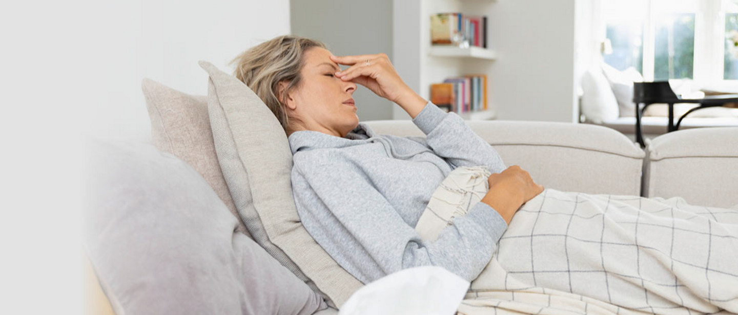 Cold during pregnancy Young woman lying on a sofa under a patterned blanket, pressing her fingers to her nose in a bright living room.