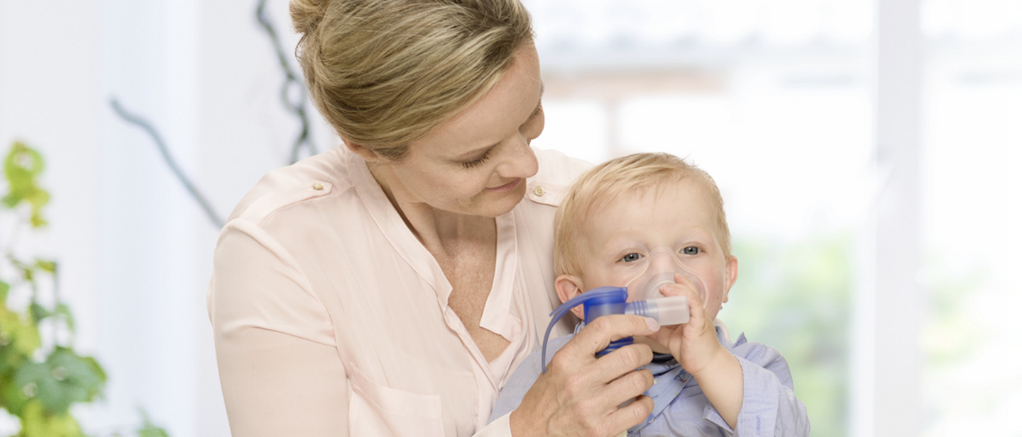 How to use a nebuliser – Using a nebuliser is easy Mother holding a small child on the lap while positioning a soft inhalation mask connected to an inhalation device.