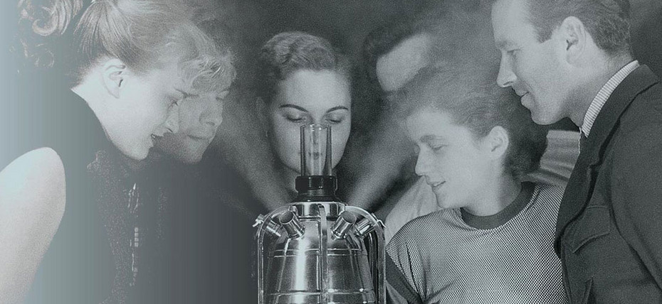 História da PARI Gruppe von Personen steht um einen Tisch und betrachtet ein Gerät, aus dem Dampf austritt.