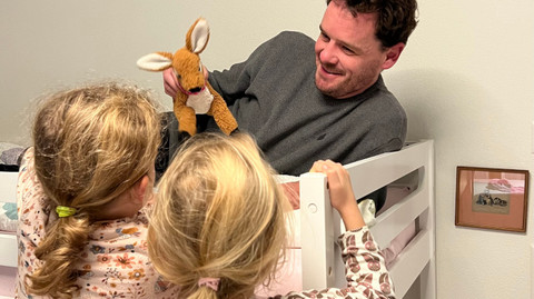 Wanting to have children: Becoming a father with cystic fibrosis? Discussion with a cystic fibrosis dad Vater liegt auf Hochbett mit Kuscheltier in der Hand, seine Töchter stehen davor und schauen ihm zu