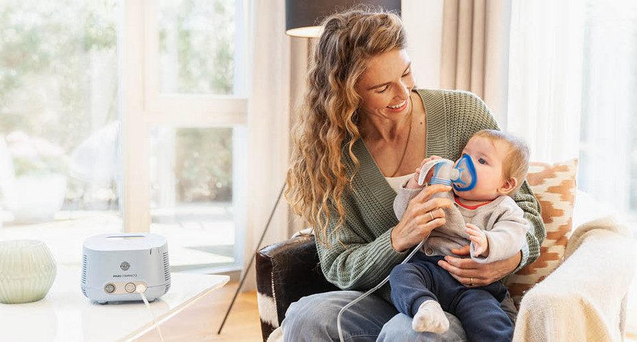 How to use a nebuliser Mother sitting in an armchair holding a baby on her lap while positioning a nebuliser at the child’s mouth.