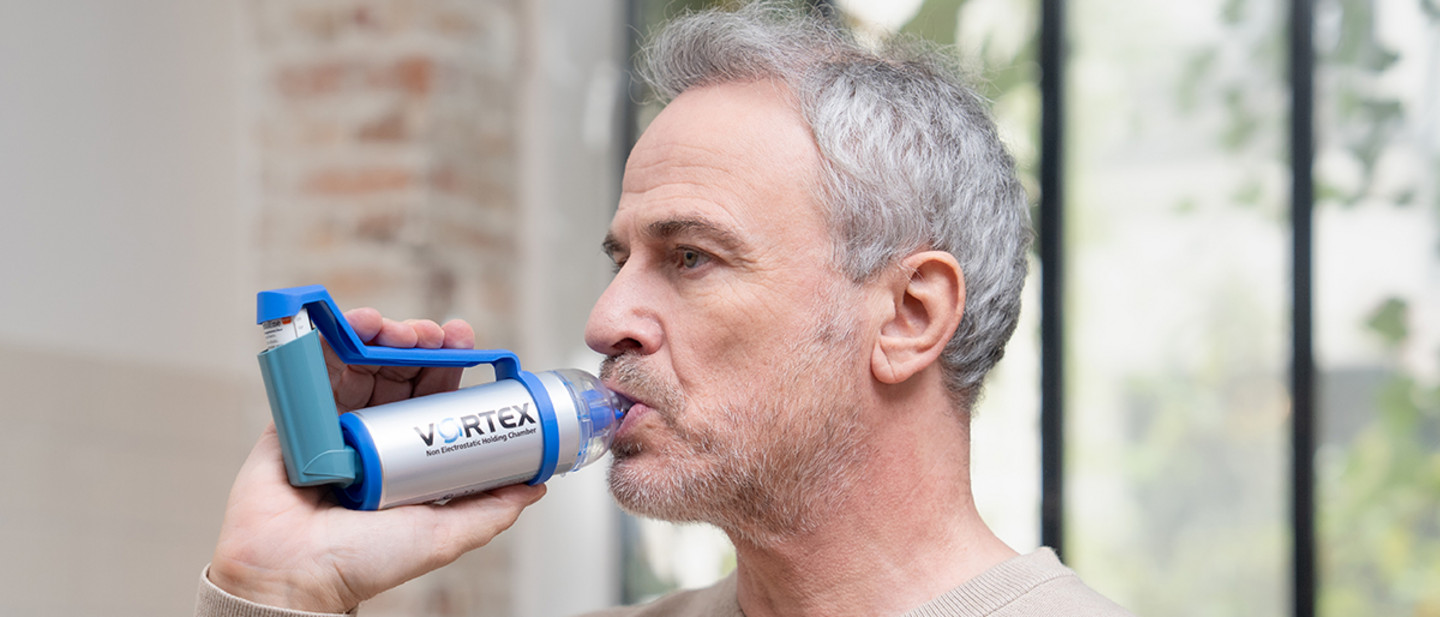Inhalation therapy with asthma spray An adult is using an asthma inhaler fitted with a VORTEX spacer in front of a window in a well-lit room.