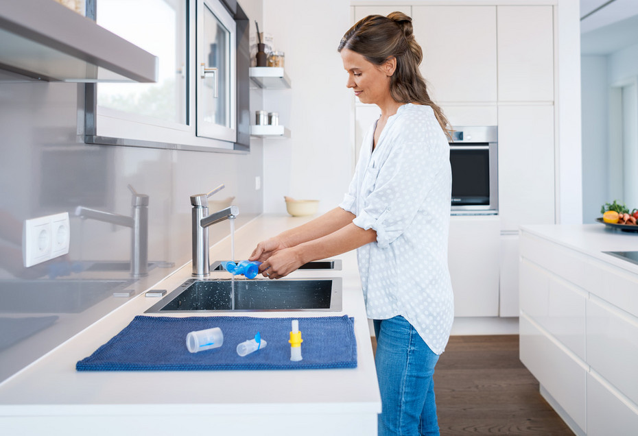 A woman stands in front of the kitchen sink, cleaning parts of the disassembled nebulizer A woman stands in front of the kitchen sink, cleaning parts of the disassembled nebulizer under running water. The already cleaned nebulizer parts lie on a cloth next to the sink to dry.