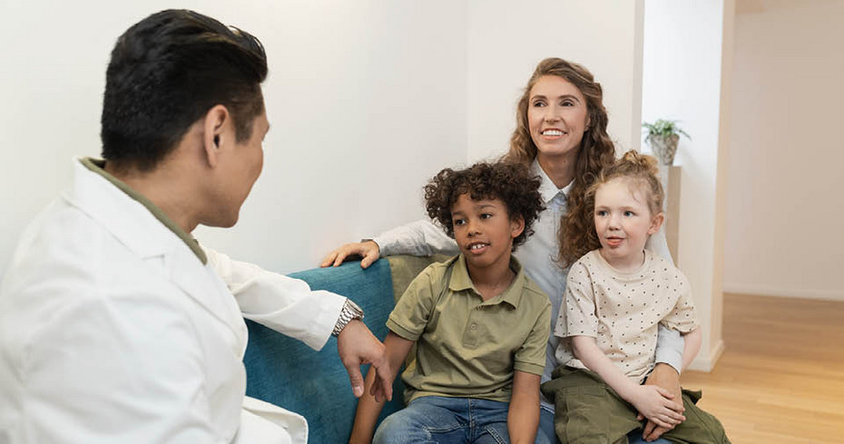 A healthcare professional talking to an adult with two children Doctor sitting on a sofa speaking with a mother and her two children seated closely together in a bright room.