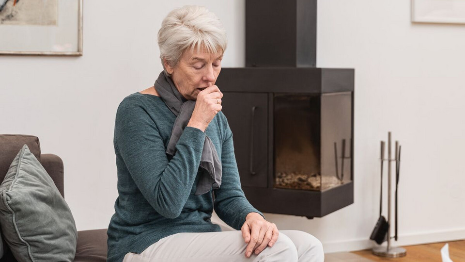 Reizhusten (trockener Husten): Ursachen und Hausmittel Ältere Frau sitzt im Wohnzimmer auf der Couch und hustet in die geschlossene Faust