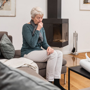 Ältere Frau sitzt im Wohnzimmer auf der Couch und hustet in die geschlossene Faust