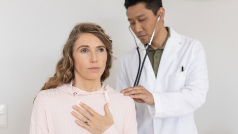 Chronic pseudomonas infestation of the lungs in a patient with cystic fibrosis: a first-hand report Junge Frau sitzt auf einer Liege mit Hand auf der Lunge, während Arzt hinter ihr steht und sie mit Stethoskop abhört