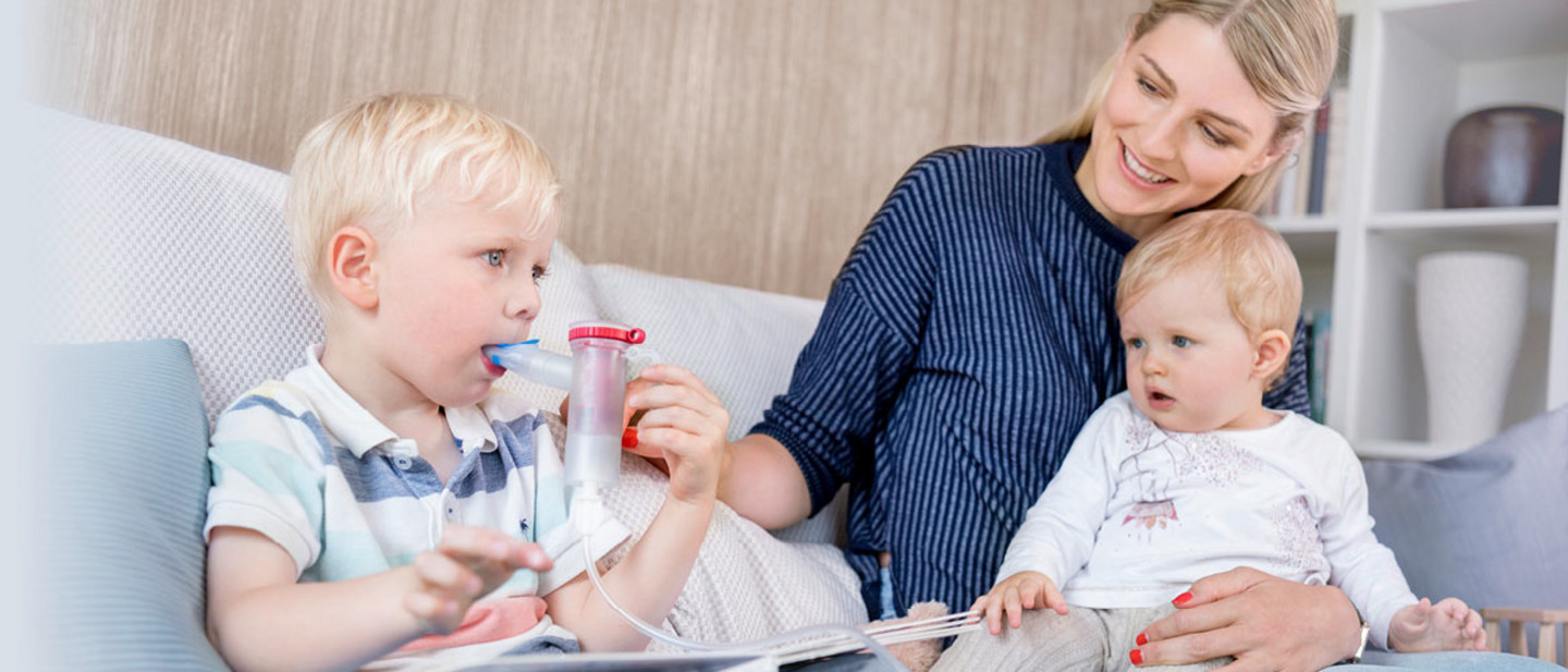 Inhalation with children Mother sitting on a sofa with two young children, helping one child hold a nebuliser while the other sits on her lap.