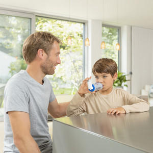 Vater und Sohn am Küchentisch, Sohn inhaliert Asthmaspray mit VORTEX Inhalierhilfe