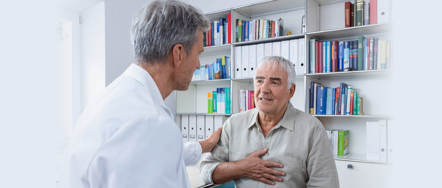 An elderly man sits in a doctor's office An elderly man sits in a doctor's office, holds his hand on his chest and talks to a doctor in a white coat who places a reassuring hand on his shoulder.