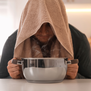 Young man sits in front of pot of steaming water and towel over his head to inhale