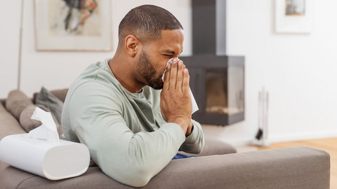 Was tun, wenn eine Allergie die Nase verstopft: Hilfreiche Tipps Junger Mann sitzt auf Couch und putzt sich die Nase