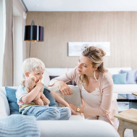 Was tun gegen Schleim im Hals beim Kind? 8 einfache Tipps für Eltern Kleiner Junge sitzt mit Kuscheltier auf der Couch, Mutter sitzt besorgt davor