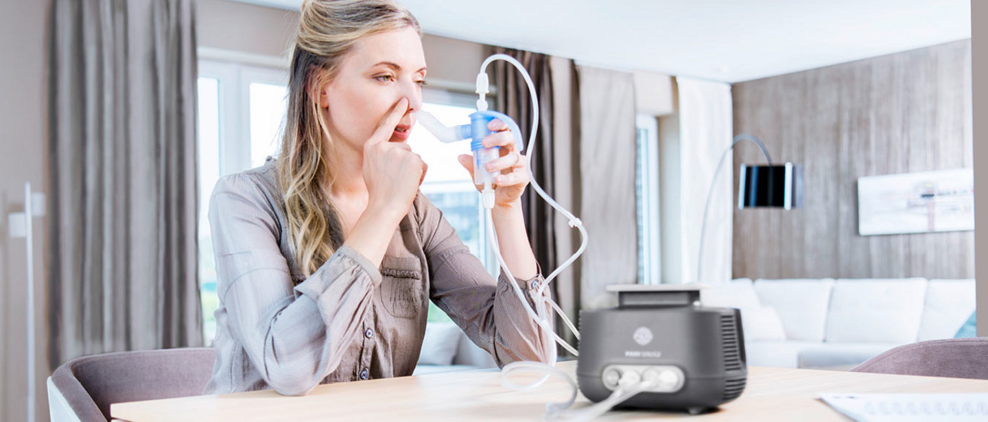 Inhalation for sinusitis Woman seated at a table holding a nebuliser connected to an inhalation device to her nose in a bright living room.