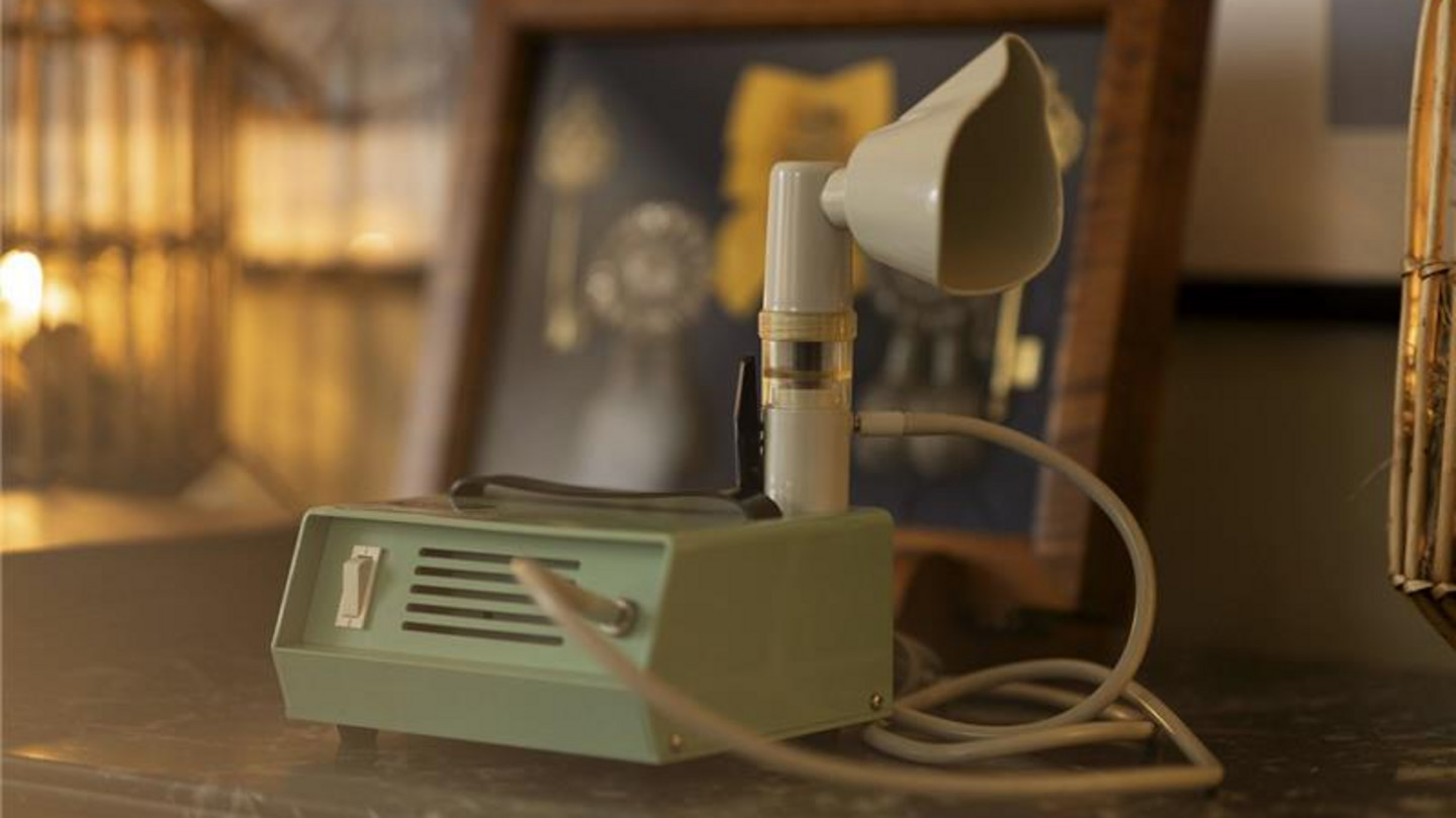 Vintage-Inhalationsgerät mit Vernebler Vintage-Inhalationsgerät mit Vernebler und Verbindungskabel auf einem Tisch, im Hintergrund ein gerahmtes Bild.