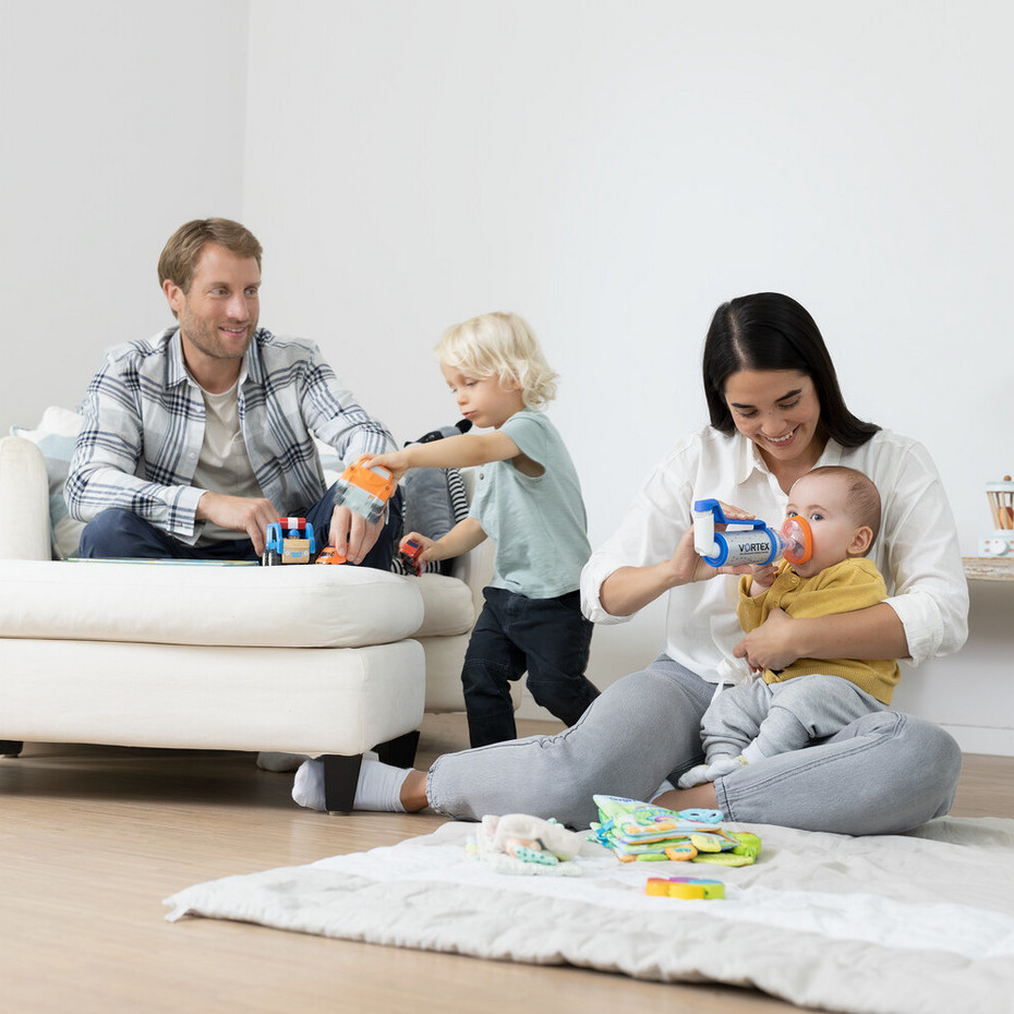 Tips to help children inhale asthma sprays: baby and child masks Young family sits in the living room, father plays with the toddler, mother sits with the baby on the floor and helps the baby to inhale asthma spray with a spacer