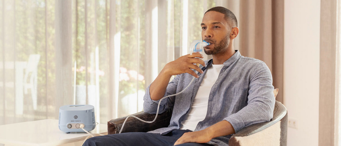 Inhalieren – das sollten sie wissen Mann mit kurzem Bart und kurzen Haaren sitzt im Sessel und nutzt einen Vernebler, der mit einem blauen Inhalationsgerät auf dem Tisch verbunden ist.