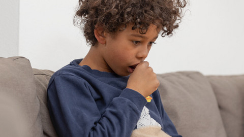 Why asthma is worse in the evening and what to do about it Junge sitzt hustend auf dem Sofa, er hält sich eine Hand vor den Mund