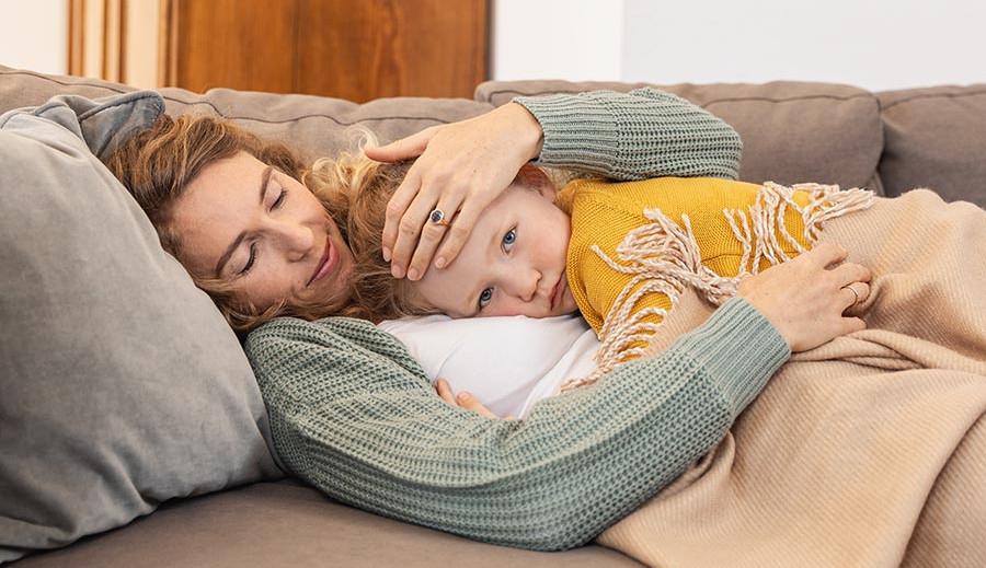 Symptoms – How the RS virus manifests itself Mother lying on a sofa holding a child close under a blanket with one hand placed on the child's forehead.