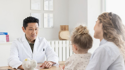Gereizte Bronchien beruhigen: Das hilft bei Bronchitis-Beschwerden Arzt sitzt auf der einen Seite seines Tisches, er unterhält sich mit einer Mutter, die ihre Tochter auf dem Schoß hat, und auf der anderen Seite des Tisches sitzt