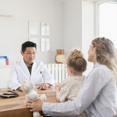 Calming irritated bronchial tubes: This helps manage bronchial symptoms Arzt sitzt auf der einen Seite seines Tisches, er unterhält sich mit einer Mutter, die ihre Tochter auf dem Schoß hat, und auf der anderen Seite des Tisches sitzt