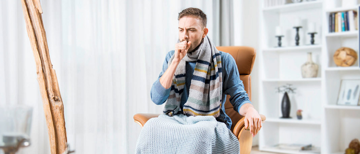Prevent infections - relieve symptoms Young man sitting in a chair with a blanket over the legs, coughing into his hand in a bright living room.