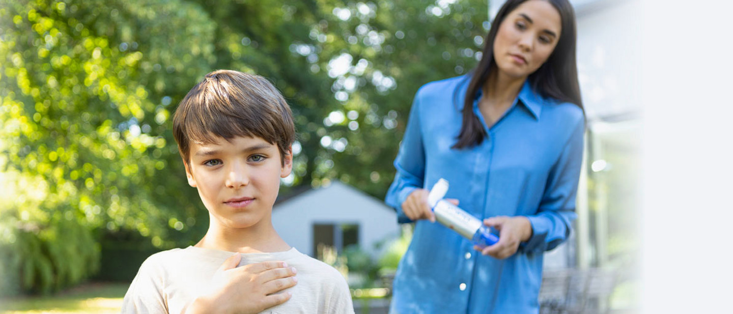 Child holds hand to chest while mother watches worriedly Boy standing in a garden with one hand on his chest, while his mother in the background holds an asthma spray.
