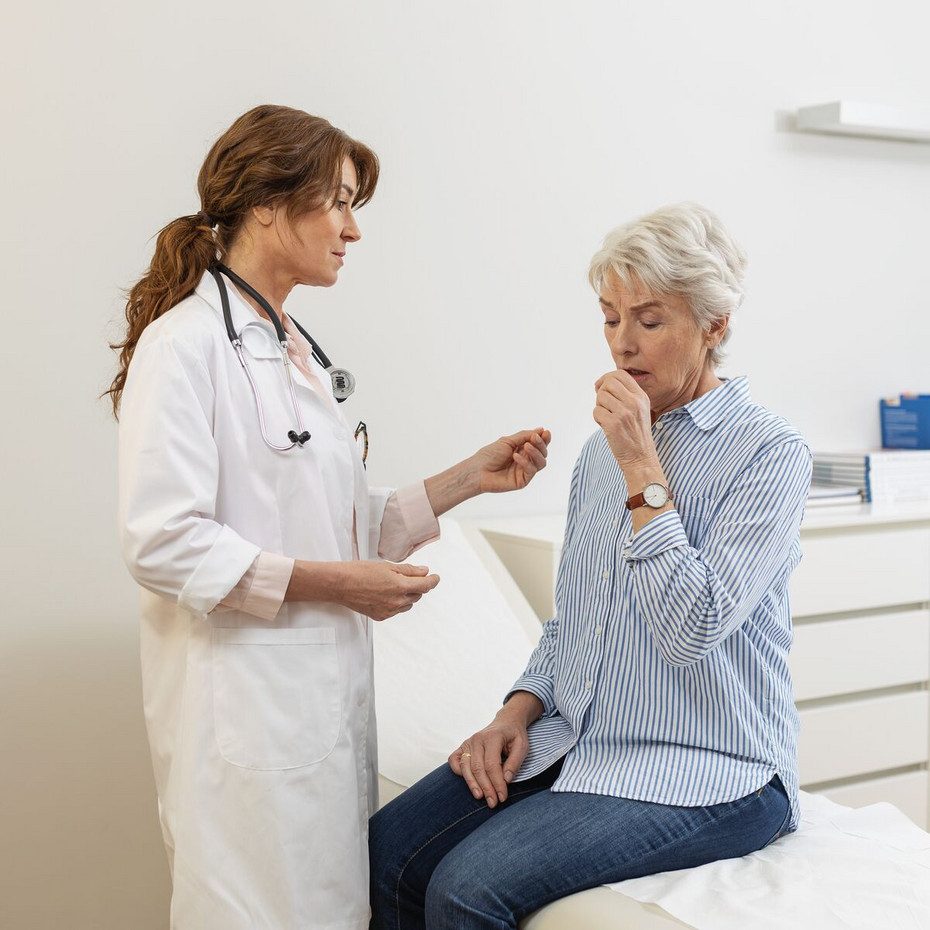 Reizhusten (trockener Husten): Wann zum Arzt? Ältere Frau sitzt auf Liege vor Ärztin mit Faust vorm Mund, die Ärztin beobachtet sie
