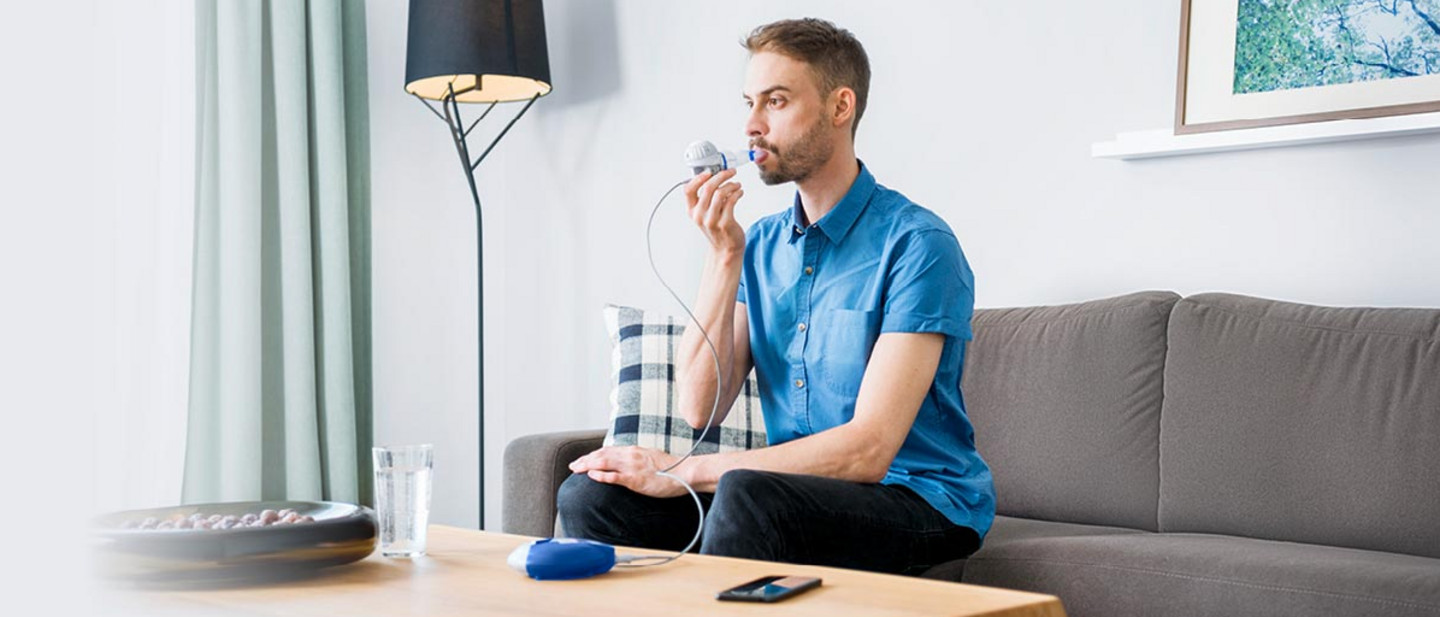 Inhalation with the eFlow®rapid Young man sitting on a sofa in a bright living room using a nebuliser connected to an inhalation device on the table.