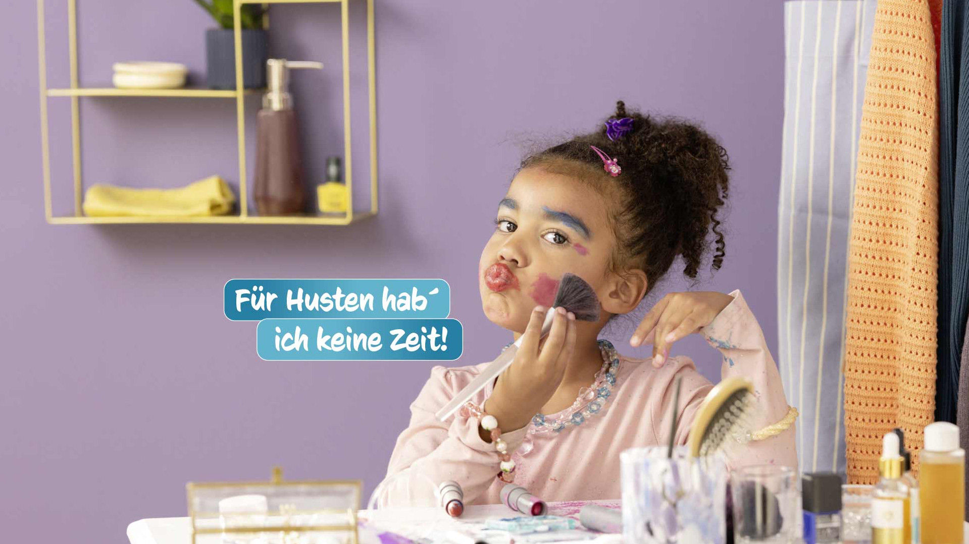 Kind sitzt vor einem Spiegel und schminkt sich spielerisch mit Make-up, während verschiedene Kosmetikartikel auf dem Tisch verteilt sind.