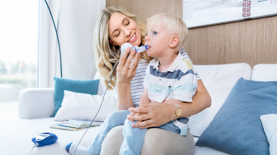 Inhalation and respiratory therapy – assistive devices for cystic fibrosis: What can you use at what age? Mother sitting on the couch with her child on her lap and helping him inhale with the eFlow rapid inhalation device for cystic fibrosis