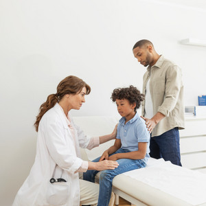 Child sits on the couch in front of a doctor, the father stands behind and has a hand on his back