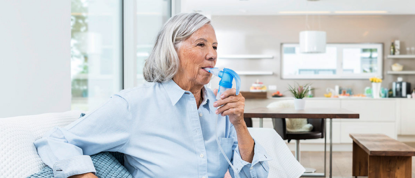 Advantages of nebuliser therapy This is how you combine physiotherapy with inhalation Older woman sitting on a sofa in a bright kitchen living area using a nebuliser with a mouthpiece.