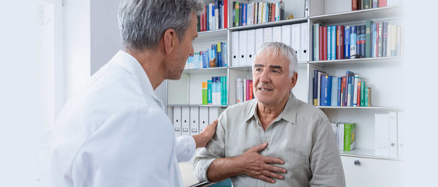 An elderly man sits in a doctor's office In a doctor's office, an older man sits with a hand on his chest while a doctor in a white coat rests a hand on his shoulder.