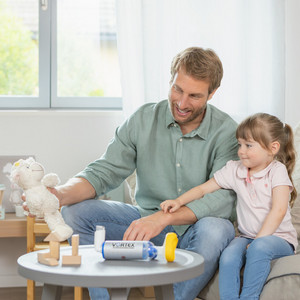 Vater sitzt mit Tochter auf Couch, er hält Kuscheltier in der Hand, das beide anschauen, auf dem Tisch vor ihnen liegt die VORTEX Inhalierhilfe