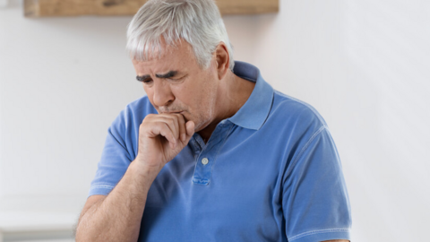 Preventing colds if you have COPD: Interview with pulmonologist Prof. Fischer Älterer Mann sitzt in der Küche am Tisch, er hustet und hält sich die Faust vor den Mund